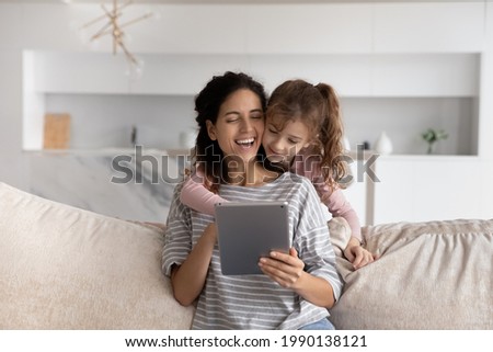Happy young Hispanic mom and small teen daughter have fun use modern tablet gadget at home together. Smiling Latino mother and little girl child talk speak on online video webcam call on pad device.