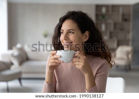 Happy millennial Latino woman drink coffee or tea from mug cup look in distance dreaming thinking of opportunities achievements. Smiling Hispanic female enjoy leisure free time at home visualizing. Royalty-Free Stock Photo #1990138127