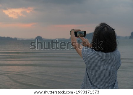 senior woman using smartphone to take photo of ocean at sunrise during summer vacation