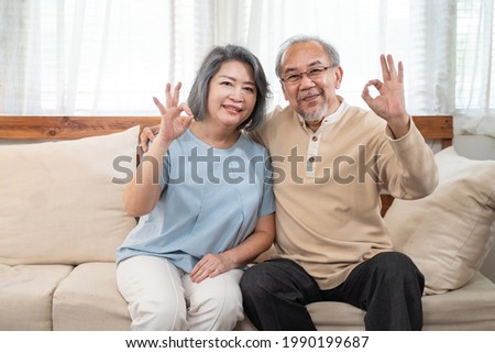 Asian happy family, Senior elderly couple enjoy retirement life at house. Older Grandfather and grandmother sit on sofa and smile, look at camera show okay gesture at home. Healthcare activity concept