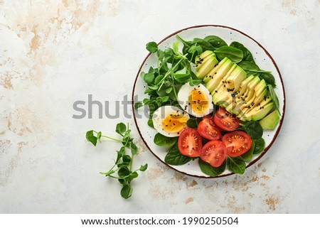 Avocado, cherry tomato, spinach and chicken egg, microgreens peas and black sesame seeds fresh salad in bowl on white stone table background. Healthy breakfast food concept. Top view.