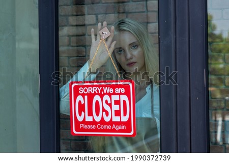 Beautiful caucasian woman with blonde hair, shop's owner standing with sad look in front of shop with closed sign, business close because of Coronavirus quarantine.
