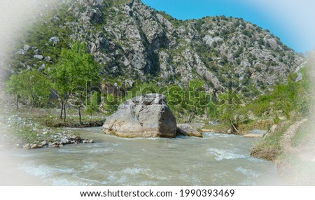 A large grey rock present on the coastline of flowing river water with large mountains behind it. Vignette effect shot of a large grey piece of rock in the water
