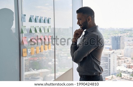 Serious thoughtful young indian businessman ceo near whiteboard with adhesive sticky notes thinking about ideas of planning strategy project standing at panoramic window of modern business office. Royalty-Free Stock Photo #1990424810