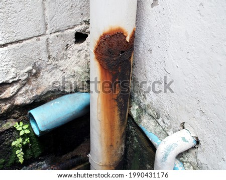 rusted and leaking white metallic water pipe, damage hole with water flow trace on joint of metal sewer pipe on outside wall corner, maintenance of home water supply systems Royalty-Free Stock Photo #1990431176