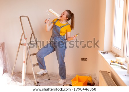 happy cute Caucasian female singing song using roller brush as a microphone. Young woman having fun during home repair and renovation Royalty-Free Stock Photo #1990446368