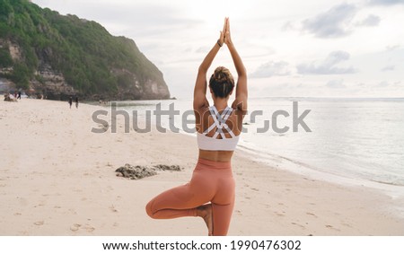 Back view of faceless young graceful female in stylish activewear standing on picturesque sandy seashore in Tree with Arms Up asana during yoga session