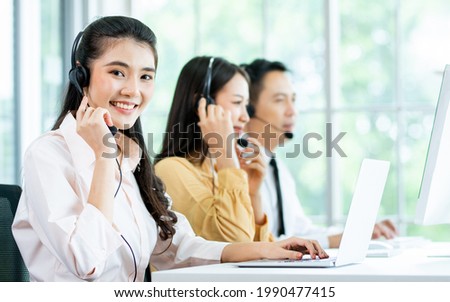 Focus Asian beautiful attractive female call center cheerful smiling with headsets, sitting, looking camera, monitoring with colleagues in office or agency at computer, giving service to customers Royalty-Free Stock Photo #1990477415