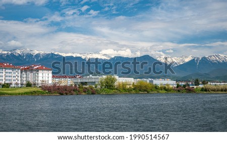Beautiful daylight scenery of snowy Caucasus mountains with a wavy lake in front and modern buildings in the middle. Sochi, Adler microdistrict, Russia. Travel destinations, hiking, leisure activities Royalty-Free Stock Photo #1990514567