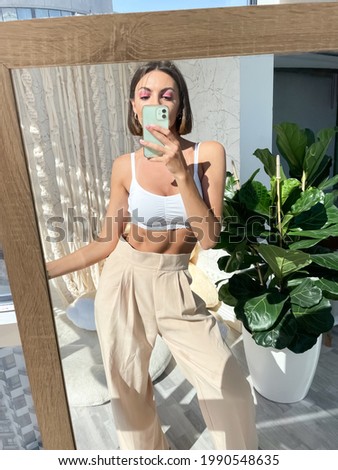 Fit tanned woman in white top and classic beige trousers at home take photo selfie on phone in mirror for social media, stories, vertical