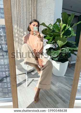 Fit tanned woman in peach sweater and classic beige trousers at home take photo selfie on phone in mirror for social media, stories, vertical