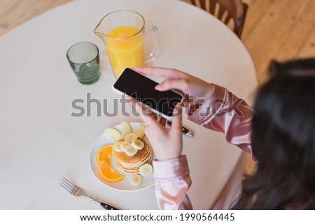 partial view of young woman holding cellphone and taking picture of pancakes with fruits near orange juice in kitchen