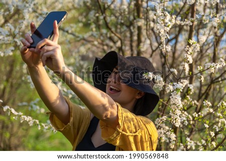 A stylish girl in a hat makes a selfie at sunset near flowering trees in the forest.