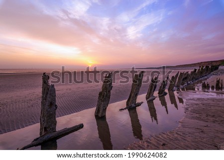 Reflections of wooden groynes on a beach with the ocean in the distance under a pink and blue sky and sun setting in the distance