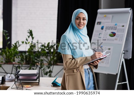 happy muslim businesswoman writing in notebook near flip chart on blurred background