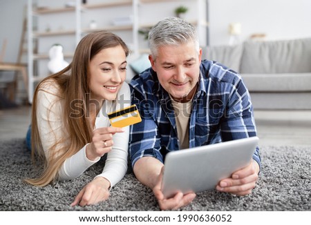 Cheerful mature couple lying on floor with tablet computer, holding credit card, making payment together in online store. Happy customers enjoying electronic store service. E-commerce concept
