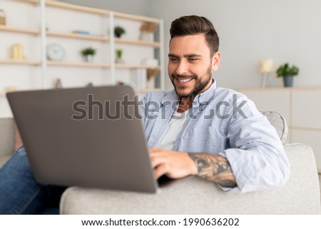 Modern job lifestyle. Happy tattooed man sitting on sofa at home and working on laptop computer distantly, surfing internet or playing online games, copy space