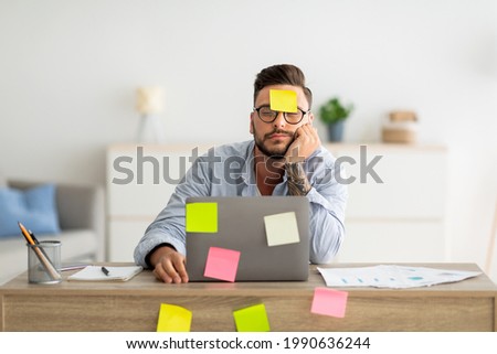 A lot of work concept. Tired freelancer man sleeping at workplace with stickers on foehead and computer, working on laptop at home office, sitting at table in living room, free space Royalty-Free Stock Photo #1990636244
