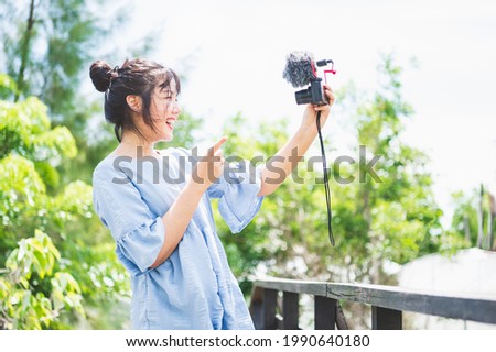 Asian woman in blue dress in public park carrying digital mirrorless camera and taking photo and vlog in happy mood. People lifestyle and leisure concept. Outdoor travel and Nature theme.