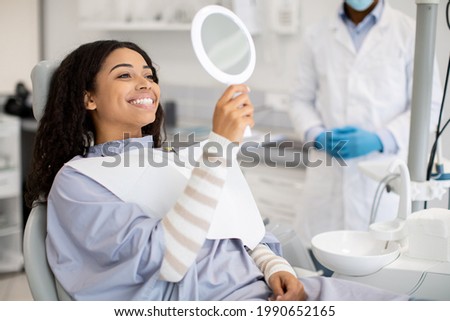 Happy Black Female Patient Looking At Mirror After Dental Treatment In Clinic, Cheerful African American Woman Sitting In Chair In Stomatological Cabinet And Enjoying Her Beautiful Smile, Closeup Royalty-Free Stock Photo #1990652165