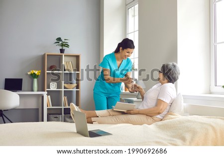 Female nurse giving glass with water to old woman patient lying on bed. Happy smiling caregiver and senior mature lady at nursing home ward. Medicine and healthcare for elderly people on retirement