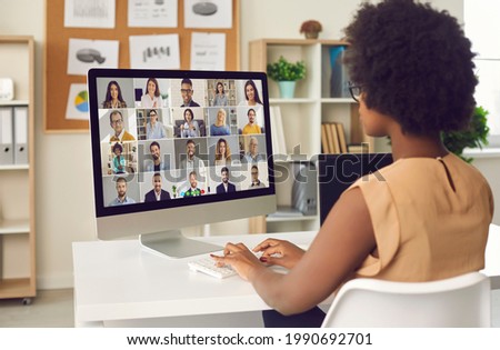 Woman having online business conference or video call staff meeting. Young black lady sitting at office desk with computer talking to team of coworkers who work from home or have hybrid work schedule Royalty-Free Stock Photo #1990692701