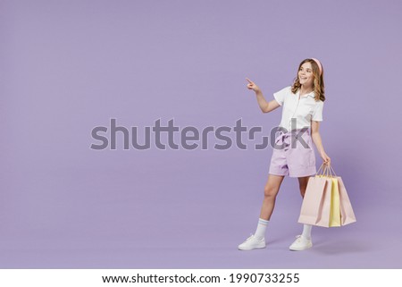 Full length little kid girl 12-13 years old in white shirt hold package purchases shopping bags point index finger aside on workspace isolated on purple background Childhood children lifestyle concept