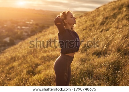 Side view of young fit female in sportswear keeping hands behind head and enjoying fresh air while resting alone after outdoor fitness workout, in hilly countryside in summer day