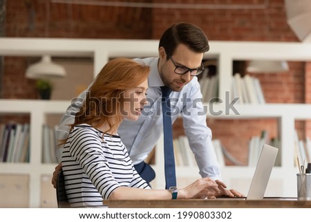 Smart confident male team leader supervisor boss helping new female employee with corporate software, working together in modern office. Happy professional managers developing online sales strategy. Royalty-Free Stock Photo #1990803302