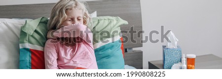 Sick ill kid girl with fever lying in bed at home with flu and sneezing in elbow. Virus cold season flu cornavirus covid-19 illness disease. Medicine and health care concept. Web banner or header. Royalty-Free Stock Photo #1990808225