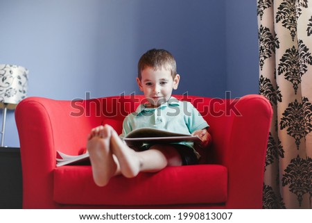 Happy Caucasian boy sitting in armchair at kids child room and reading book. Early age kid child development and literacy education. Candid home authentic childhood lifestyle concept.
