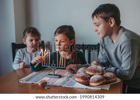 Brother and sisters siblings lighting candles on menorah for Jewish Hanukkah holiday at home. Children celebrating Chanukah festival of lights. Dreidel and Sufganiyot donuts in plate on a table. 