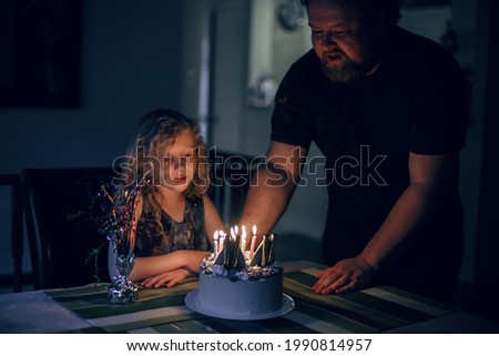 Cute funny blonde girl blowing candles on birthday cake in dark room. Happy birthday party celebration. Making a wish while blowing candles tradition. Child celebrating birthday at home.