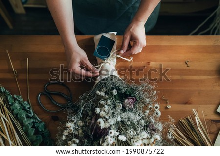 Hands of Female Florist Making Everlasting Bouquet of Dried Flowers at Wooden Table at her Flower Shop.
Close up of unrecognizable woman entrepreneur arranging bouquet of a dry flowers and plants.