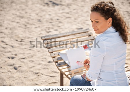 Beautiful Hispanic woman looking away and drawing using watercolors sitting on a wooden chaise lounge on the beach sand