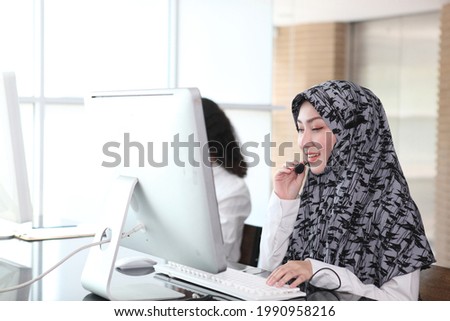 Asian Muslim female  with headsets working in a call centre,Call center agent with headset working on support hotline in modern office. Call center worker accompanied by her team. speaking to partner
