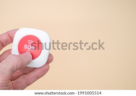 An elderly hand holding a wireless SOS emergency alarm button isolated on light brown background