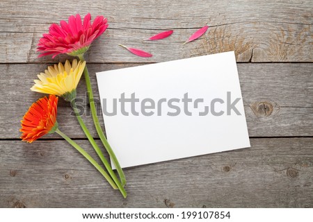 Three colorful gerbera flowers and blank gift card or photo frame on wooden table