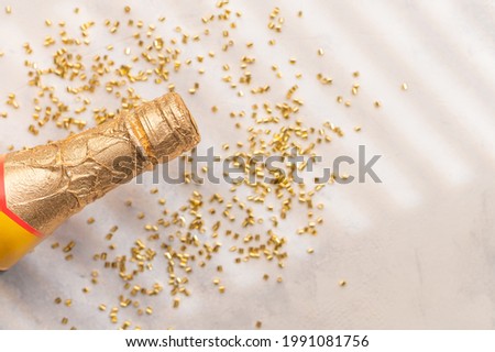 On a white background, we see in the left corner of the photo a bottle of champagne with a golden neck. Confetti is scattered around. White and gold tones. Background. Texture. Wallpaper.