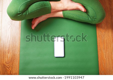 A woman watches yoga classes on her smartphone while sitting on a green exercise mat. The concept of training in the living room of the house. Technology and sports
