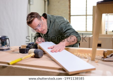 carpenter checking the wooden material, thinks if there are irregularities on it, wearing safety goggles for woodworking, looking seriously, concentrated on work. crafts, woodworking, carpentry Royalty-Free Stock Photo #1991138585