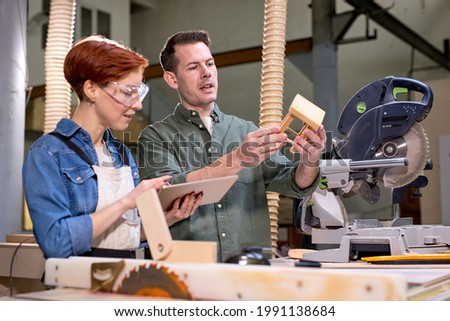 Joiners co-workers work on making wooden chair stool, engaged in woodworking, carpentry together. Man holding small copy of chair in hands, having talk, sharing ideas and opinions, cooperating