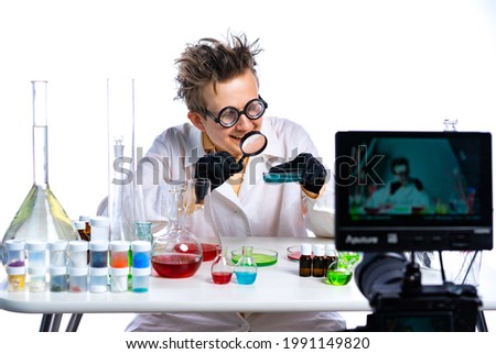 A disheveled doctor in glasses with large lenses sits at a table with multi-colored flasks. the camera is recording video. funny scientist - blogger. home video. on-line translation. remote learning