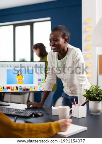 Satisfied african photo editor discussing with colleague how retouching image, working on PC. Woman retoucher editing assets in pro software app with graphic tablet sitting in creative agency