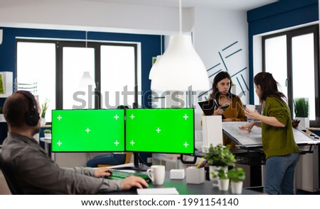 Employee with headphones using dual monitror setup with green screen, chroma key mock up isolated display sitting in video production studio. Man ditor processing film montage on pc in creative agency
