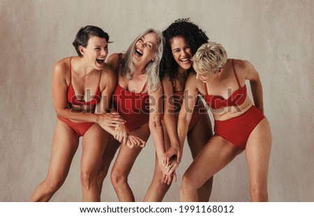 Natural women of all ages celebrating their ageing bodies. Four happy and body positive women having fun while wearing red swimwear against a studio background. Royalty-Free Stock Photo #1991168012