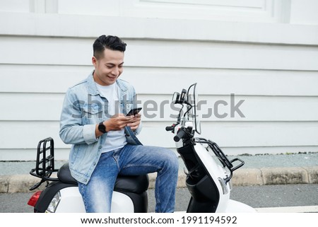 Smiling young Vietnamese moped driver in denim outfit sitting on modern bike and using smartphone while communicating online
