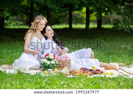 Two girls are resting in park sitting on a picnic blanket. Friends is making picnic outdoor with fruits and wine. Blonde and brunette girl in nature.