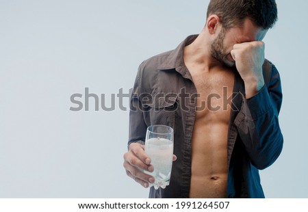 Unhealthy man with headache hold glass with pill dissolving. Young bearded guy touch face. Isolated on turquoise background. Studio shoot. Copy space