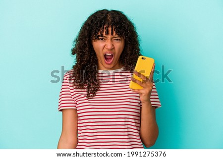 Young mixed race woman holding a mobile phone isolated on blue background screaming very angry and aggressive.